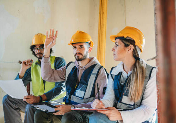 Three people are listening to their foreman on a construction site. They are sitting on the worksite.